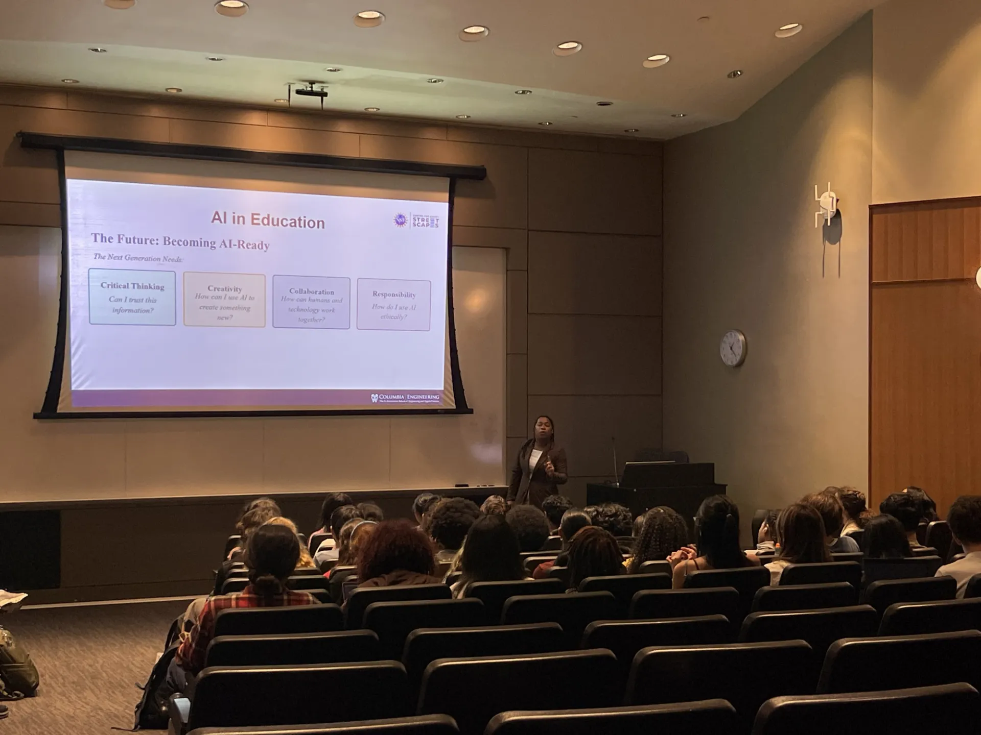 A presentation to a group of high school researchers in an auditorium in front of a large projector.